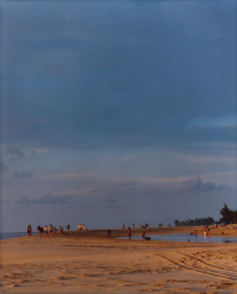 Beach scene at dusk features a group of people on the shoreline with sand and water in the foreground and a cloudy sky above.