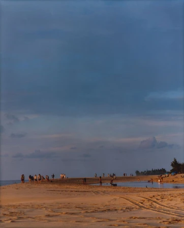 Beach scene at dusk features a group of people on the shoreline with sand and water in the foreground and a cloudy sky above.