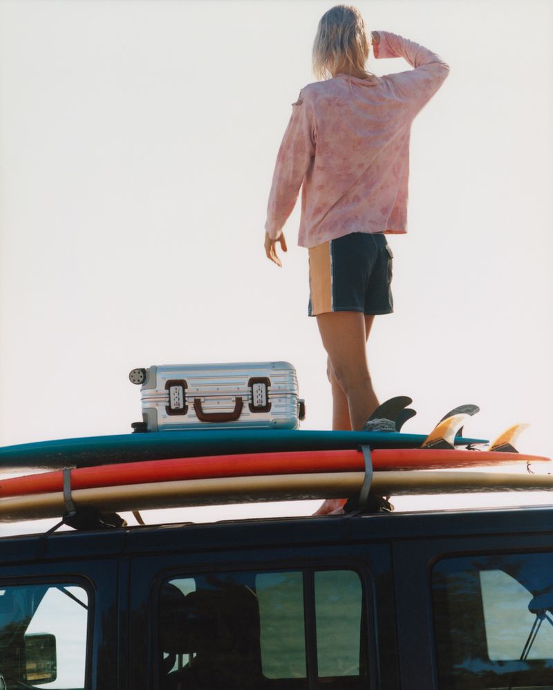 Person standing on a vehicle roof rack next to luggage and surfboards, looking out towards the horizon in a sunny, outdoor setting.