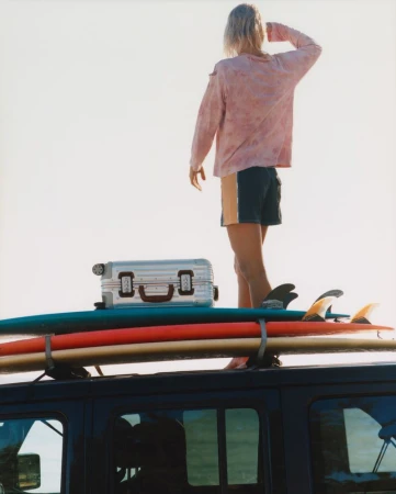 Person standing on a vehicle roof rack next to luggage and surfboards, looking out towards the horizon in a sunny, outdoor setting.