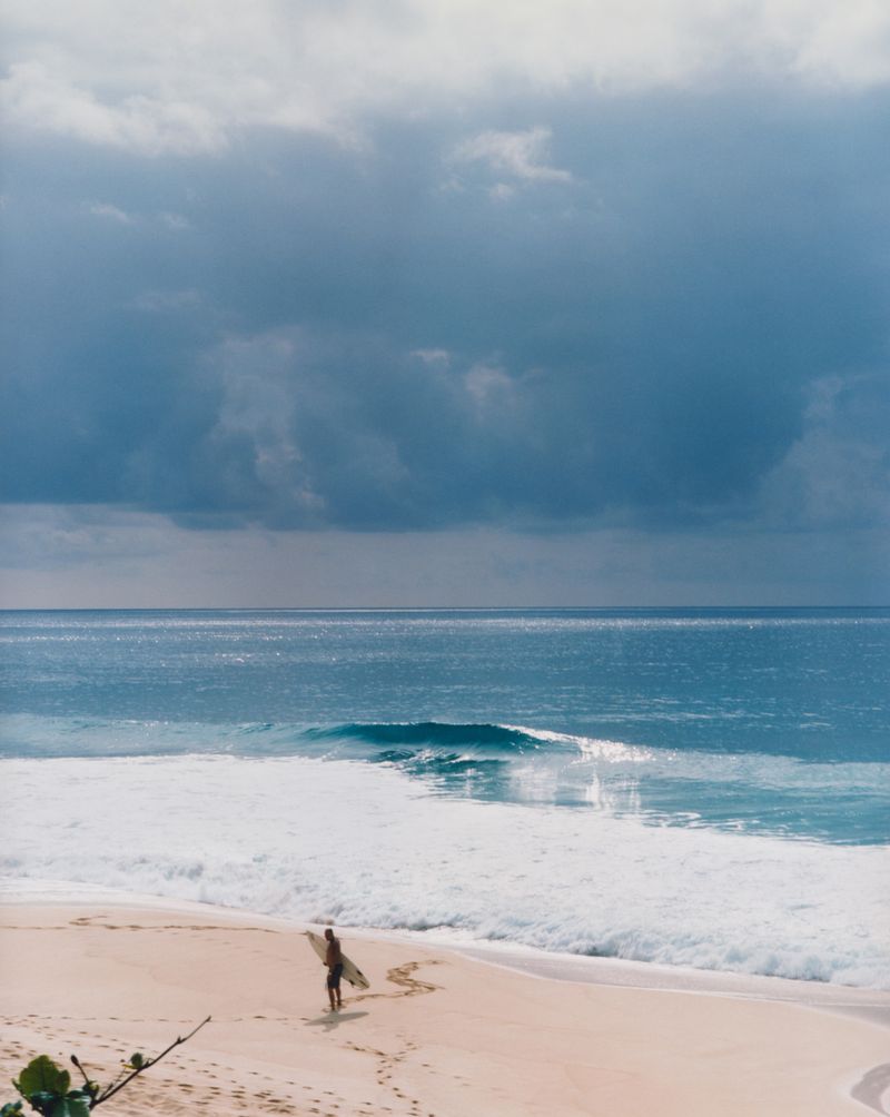 A solitary surfer walks along a sandy beach carrying a surfboard towards the ocean under a cloudy sky.