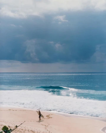 A solitary surfer walks along a sandy beach carrying a surfboard towards the ocean under a cloudy sky.