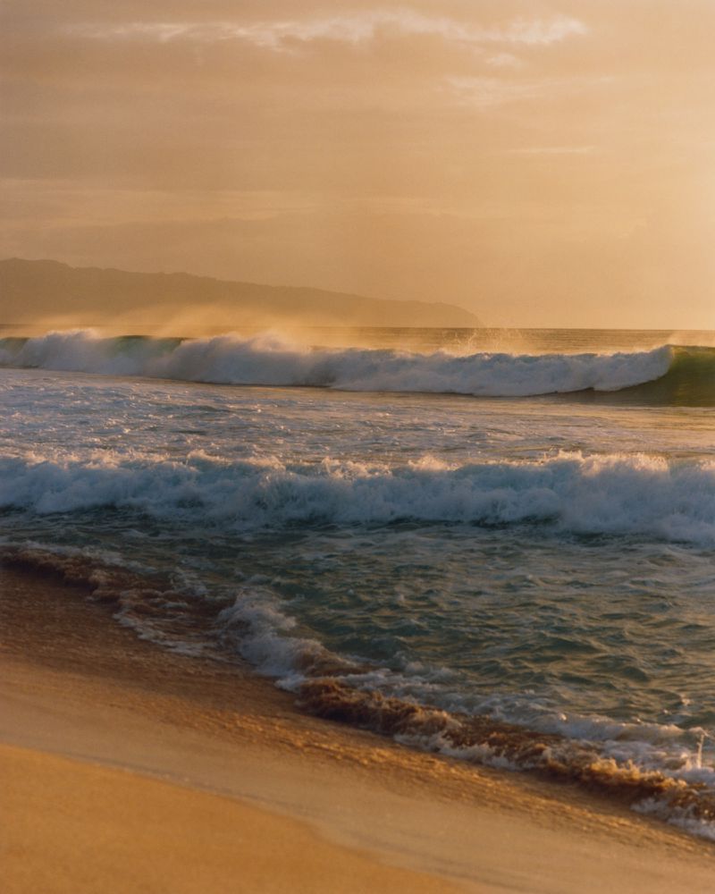 Ocean waves crashing on a sandy beach, lit by a golden sky at sunrise or sunset, with distant mountains adding to the scenic view.