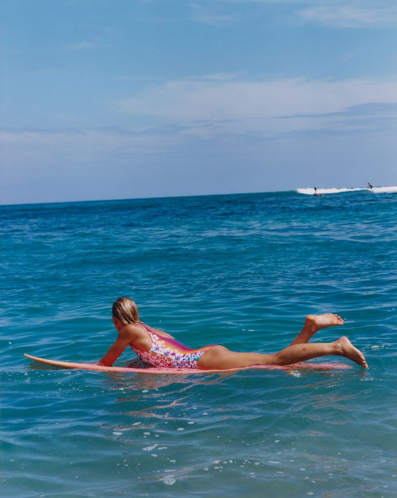Young woman in floral swimsuit lying prone on surfboard in blue ocean water, blue sky day. Surfers in distance.