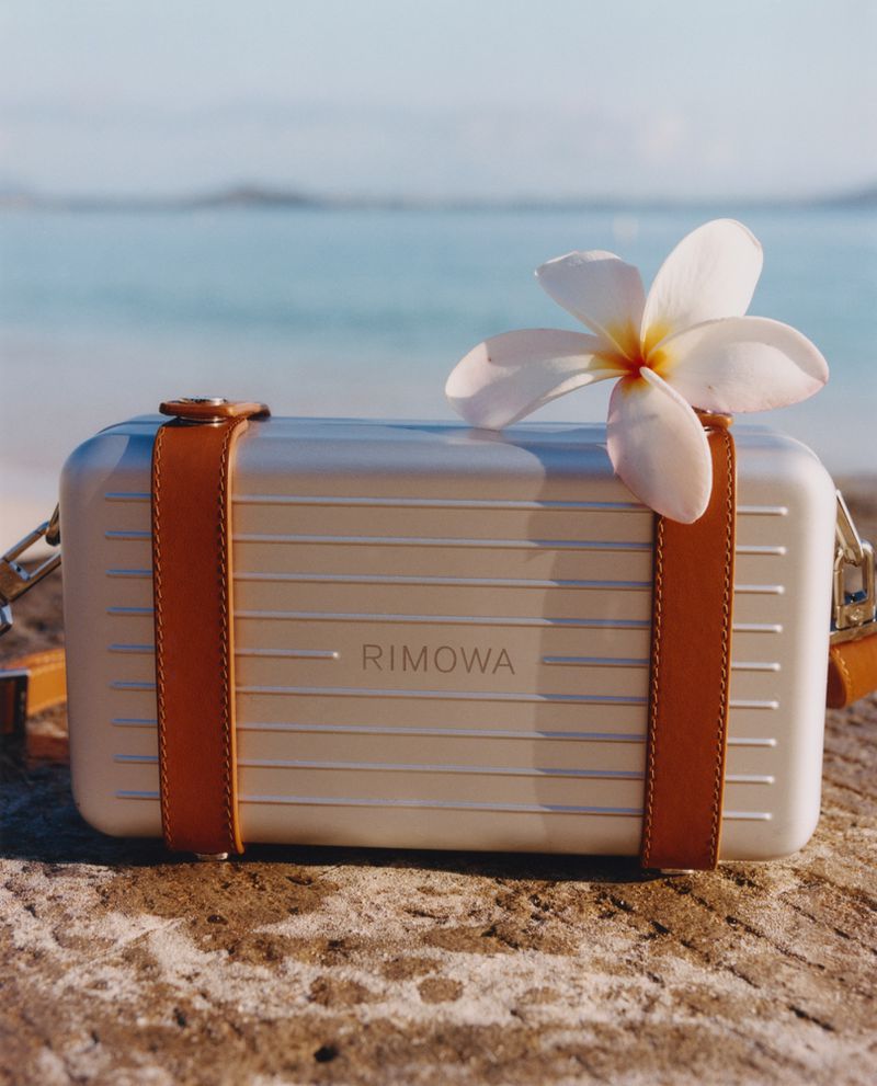 Rimowa handbag displayed on a sandy surface with a white tropical flower, set against an out-of-focus beach and ocean backdrop.