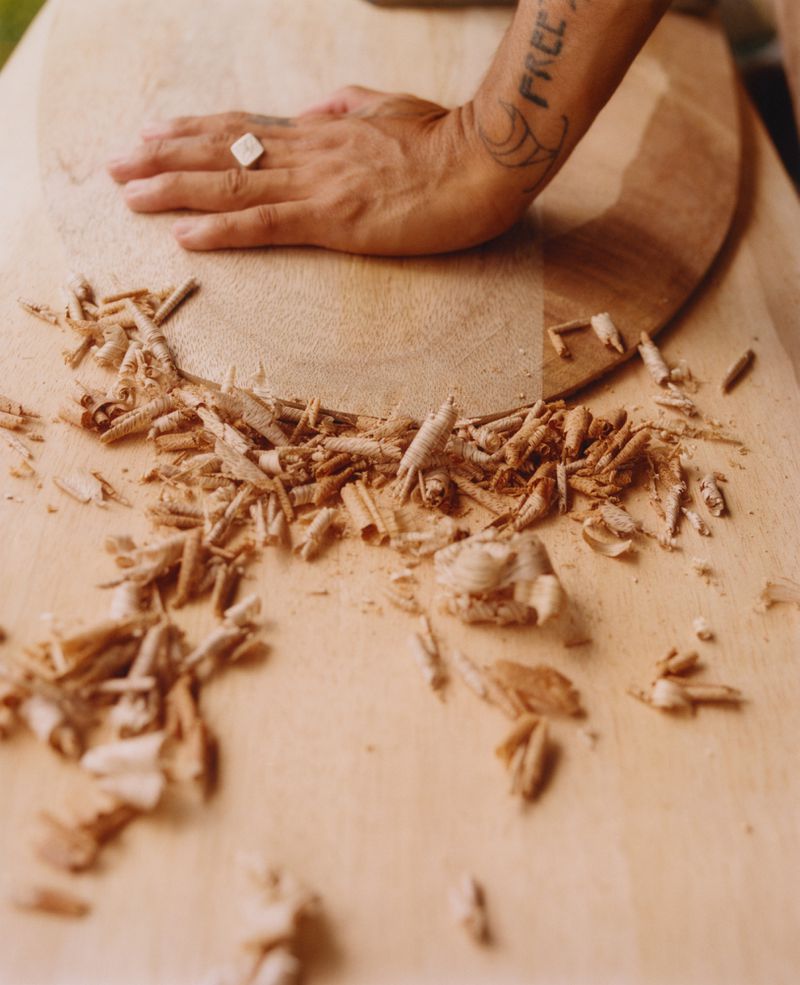 Hand rests on wooden surface with scattered wood shavings, tattoos visible on arm, focus on texture and woodworking process.