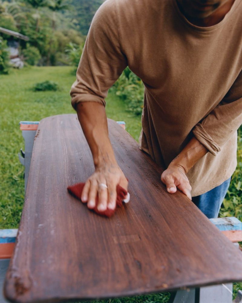 Person refinishing a wooden board with a red cloth outdoors. Woodworking project. Man sanding a long board on a table in a yard.