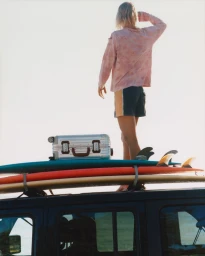 Person standing on a car roof rack beside stacked surfboards and a silver RIMOWA suitcase against a bright open sky.