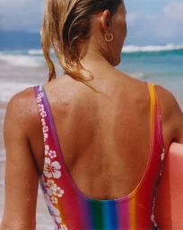 Close-up back view of a woman in a colorful floral swimsuit holding a surfboard near the ocean.