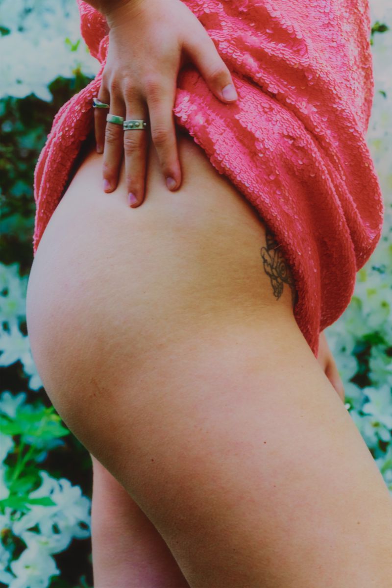 Close-up of an adult female in a short, sparkly, pink dress with a hand resting on her thigh, set against a blurred background of flowers.
