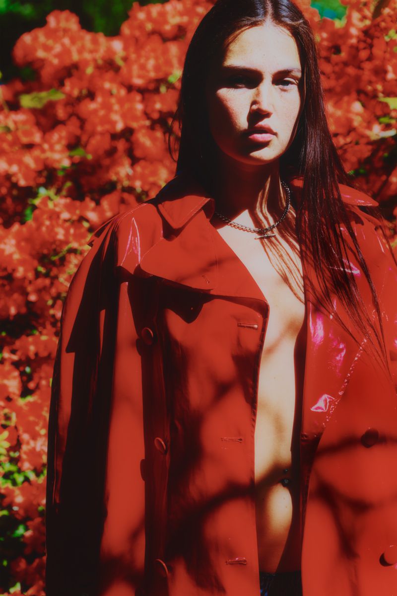 A young woman with long brown hair poses in an unbuttoned red trench coat in front of leafy foliage, shot in an outdoor setting.
