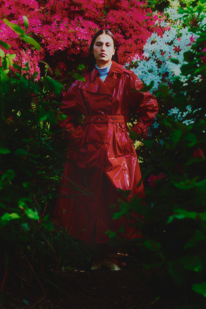 A woman in a shiny red trench coat stands among lush green foliage and colorful pink, white, and red flowers in an outdoor setting.