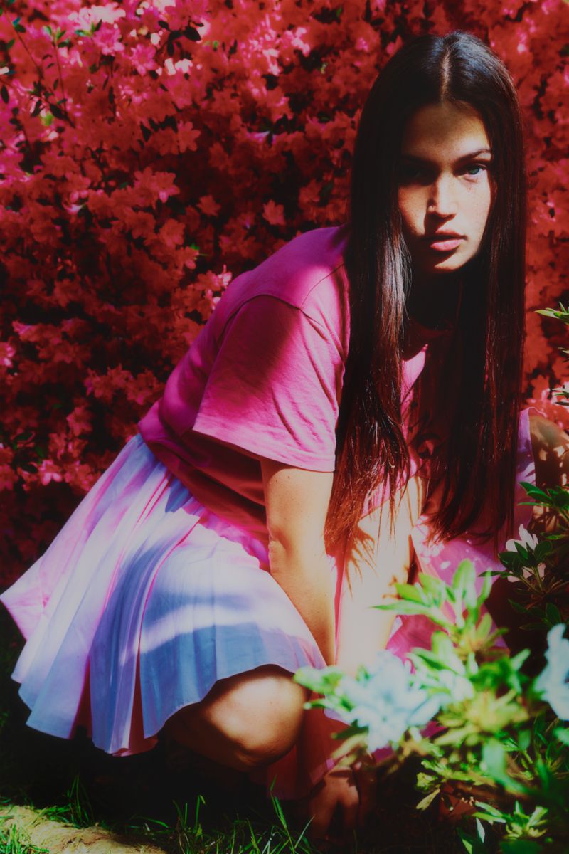 Portrait of a woman in a pink top and pleated skirt crouching in a garden with pink flowers and green foliage in soft lighting.