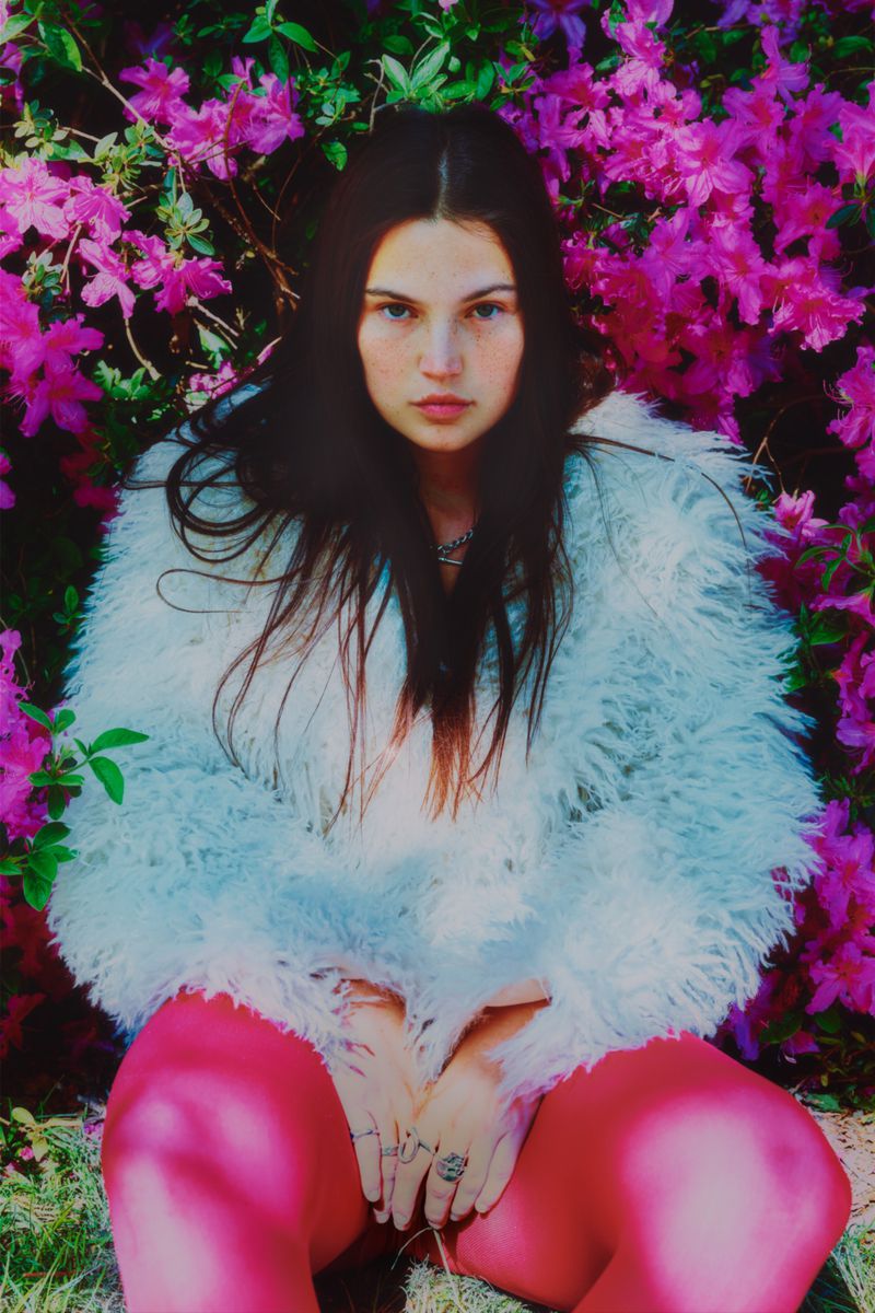 A young woman with long brown hair poses in a white faux-fur coat and pink tights, seated in front of a backdrop of pink flowers and green leaves.