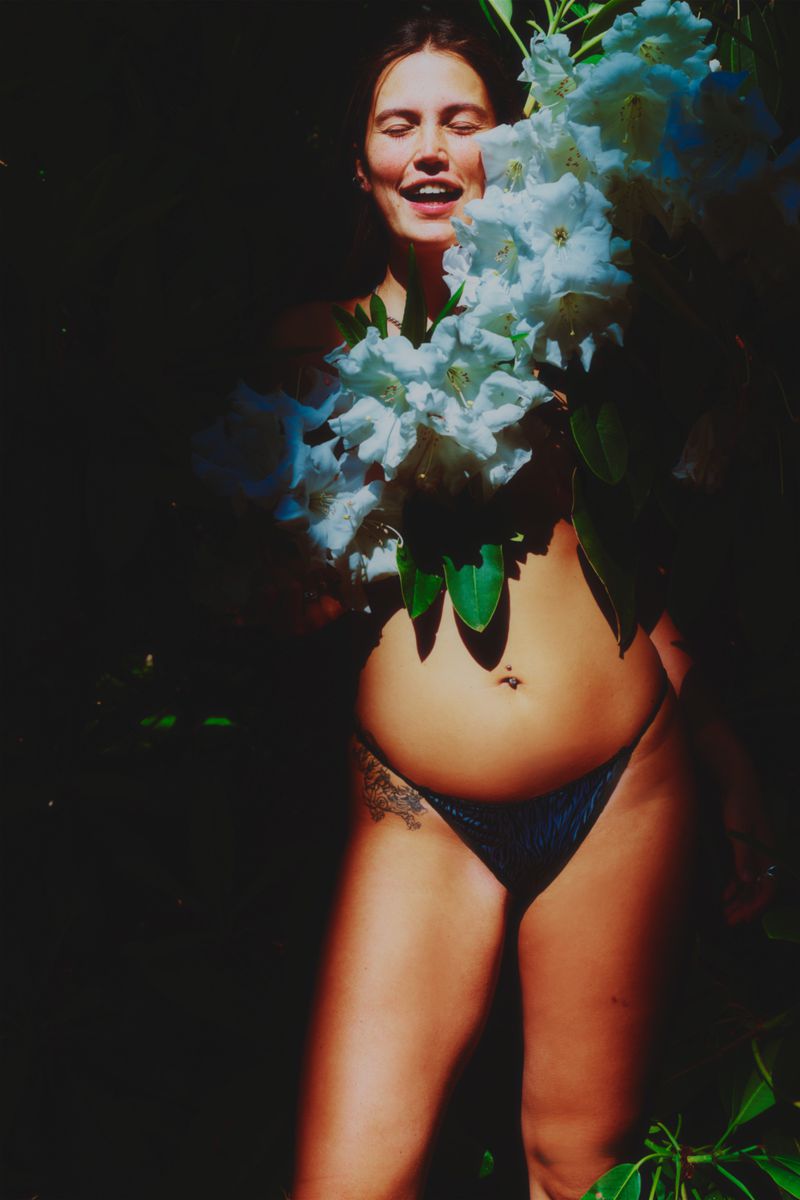 Woman in a bikini poses with white flowers in a dark outdoor setting, smiling slightly, with a tattoo visible on her hip.