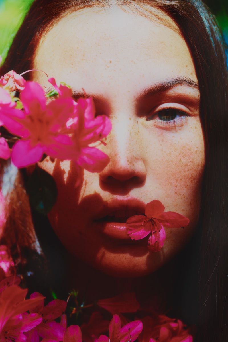 Close-up portrait of a brunette woman with freckles, accentuated by vibrant pink flowers framing her face in an outdoor setting.