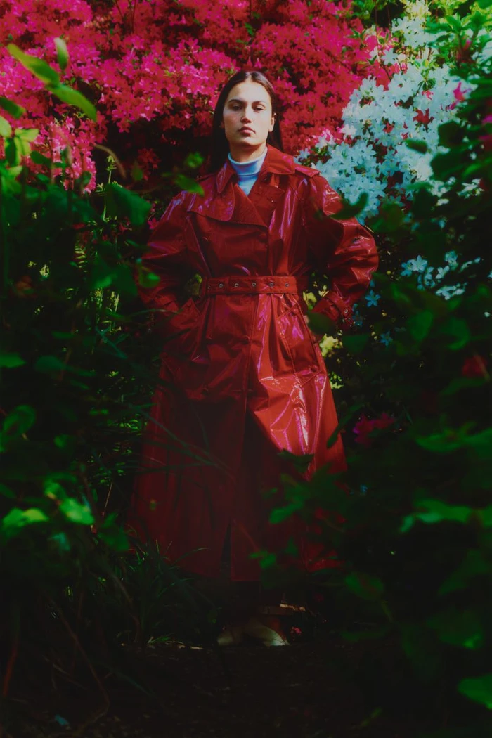 A woman standing among lush blooming pink and white garden flowers wearing a glossy red belted trench coat.