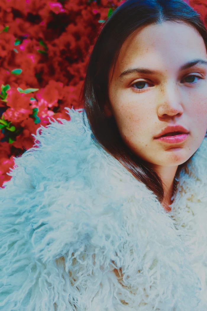 Close-up portrait of a young woman with dark hair wearing a fluffy white textured coat against a vibrant red floral backdrop.