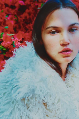 Close-up portrait of a young woman with dark hair wearing a fluffy white textured coat against a vibrant red floral backdrop.