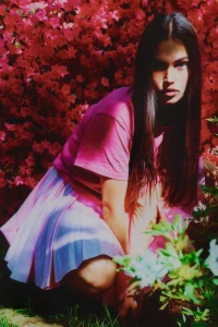 Young woman in a pink t-shirt and light skirt squatting in front of a vibrant bush of pink flowers