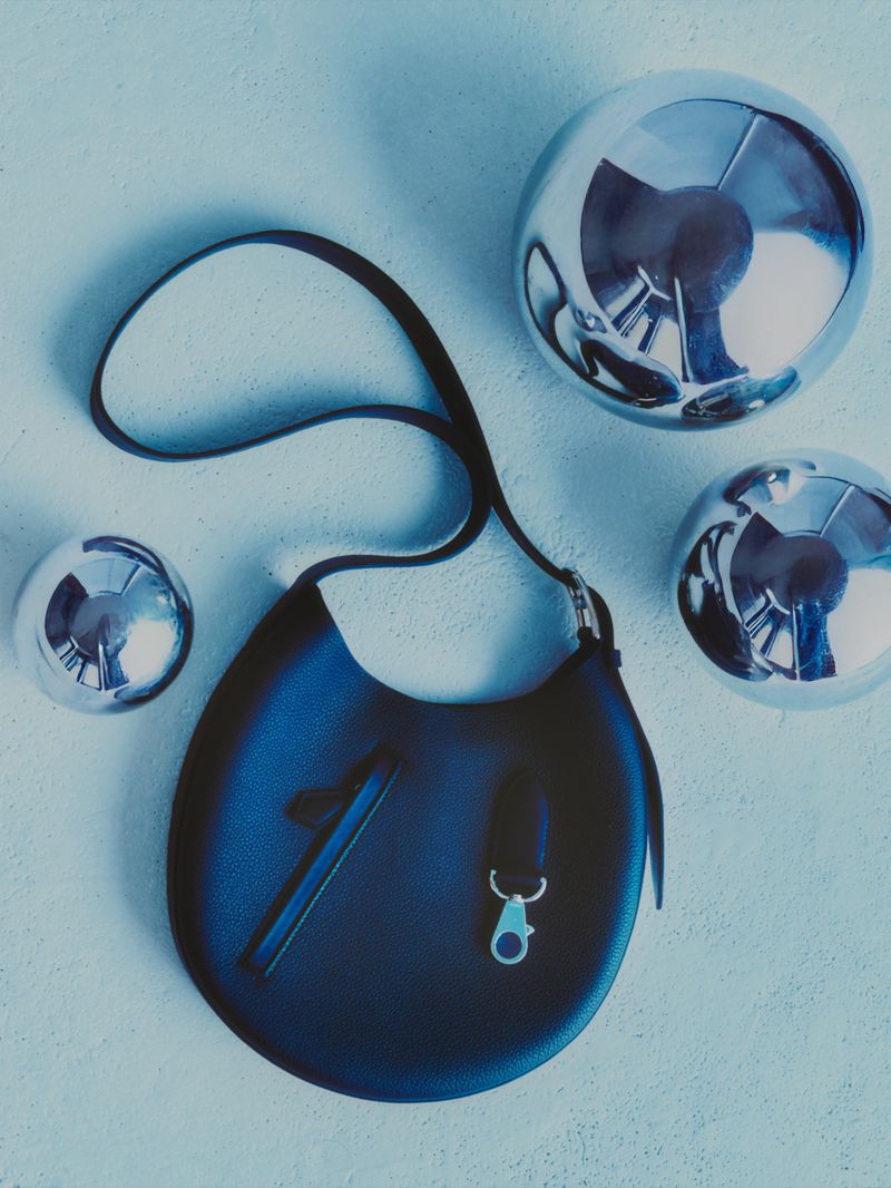 Black leather handbag with a strap, suspended against a textured blue backdrop, surrounded by three metallic silver spheres.