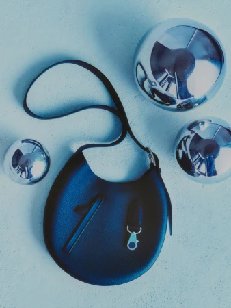 Black leather handbag with a strap, suspended against a textured blue backdrop, surrounded by three metallic silver spheres.