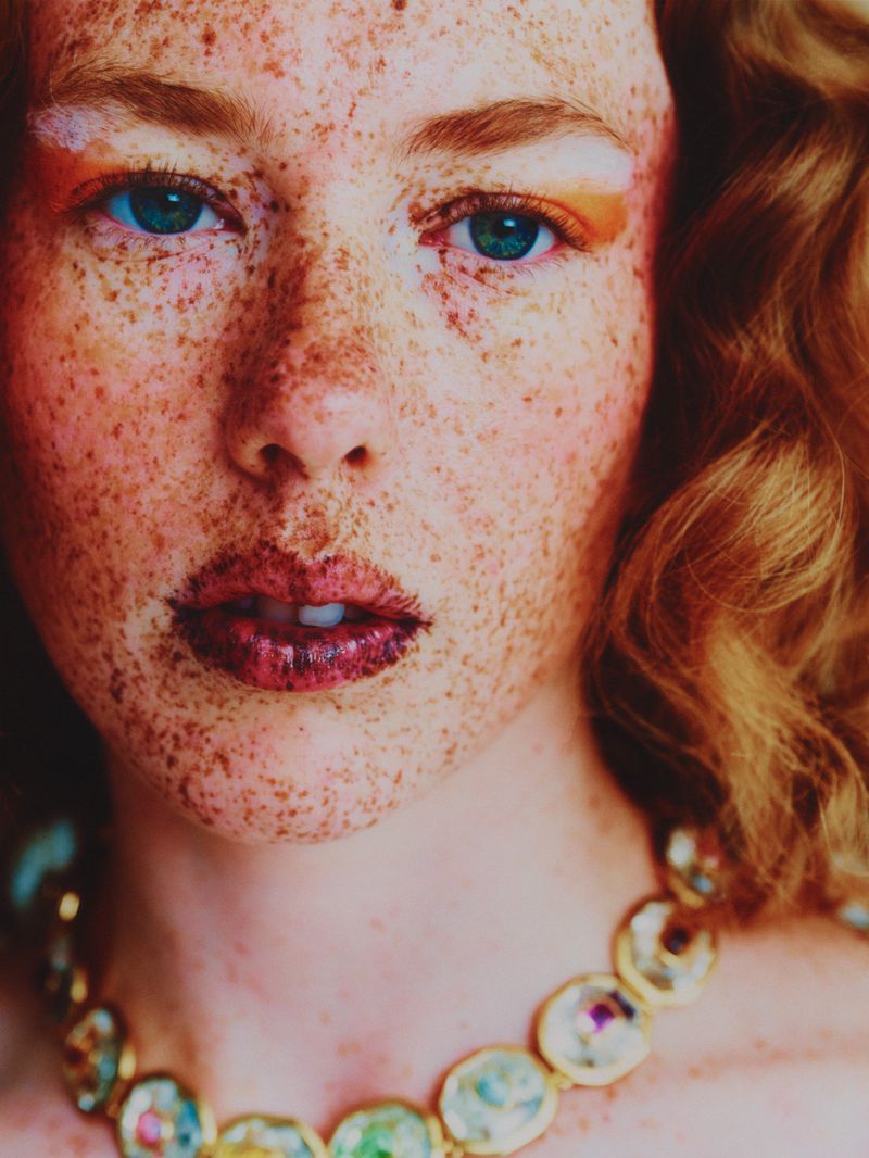 Close-up portrait of a red-haired woman with freckles, wearing a jeweled necklace, with eye-catching orange makeup around her eyes.