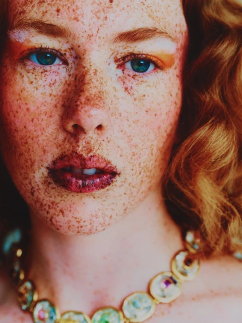 Close-up portrait of a red-haired woman with freckles, wearing a jeweled necklace, with eye-catching orange makeup around her eyes.