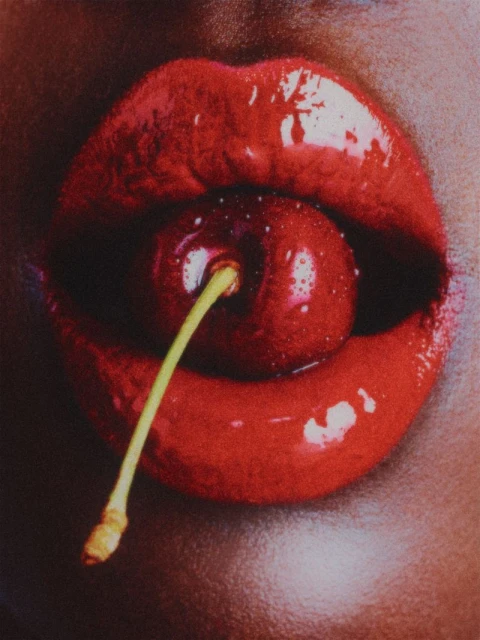 Close-up of a person holding a cherry in their mouth, featuring red lipstick and visible skin texture against a dark, indistinct backdrop.