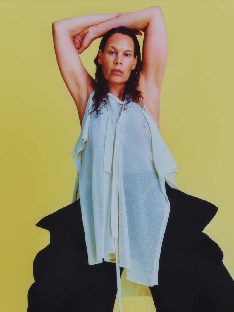Woman in oversized black pants and a light green sleeveless top poses in front of a solid yellow background in a studio shot.