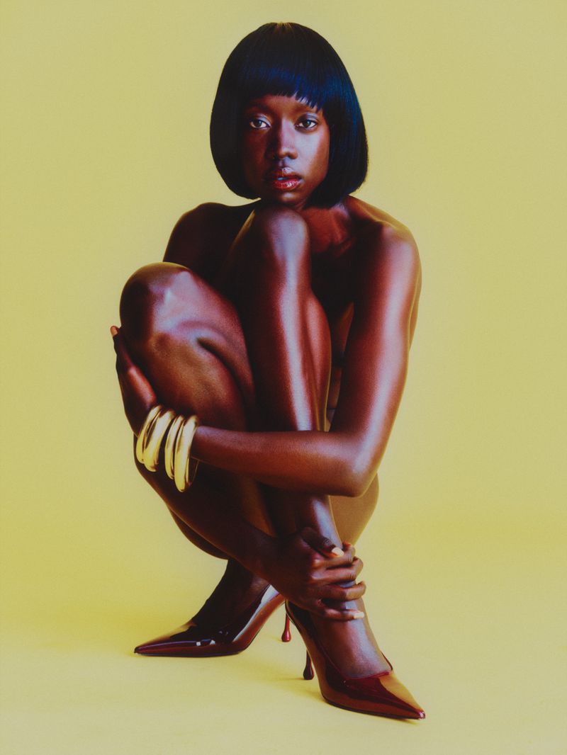 Portrait of an African American woman with short hair wearing heels, sitting in a studio against a light-yellow background.