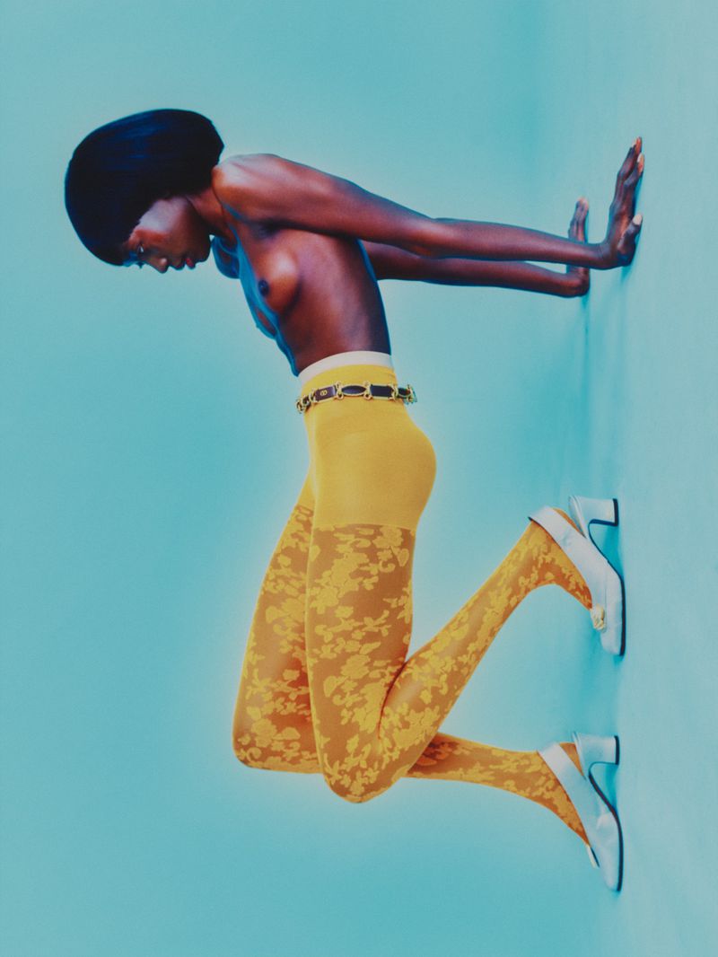 Dark-skinned woman strikes a pose on a light blue backdrop, highlighting her body and fashion choices with floral patterned tights.