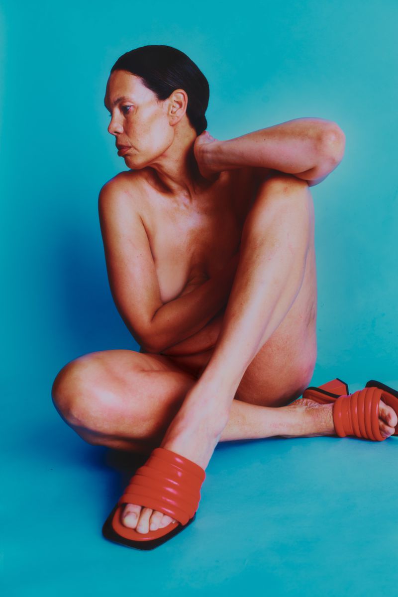 A topless adult woman sits cross-legged with red sandals on a blue background, gazing leftward with her hand at her neck.