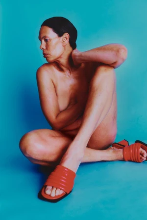 A topless adult woman sits cross-legged with red sandals on a blue background, gazing leftward with her hand at her neck.