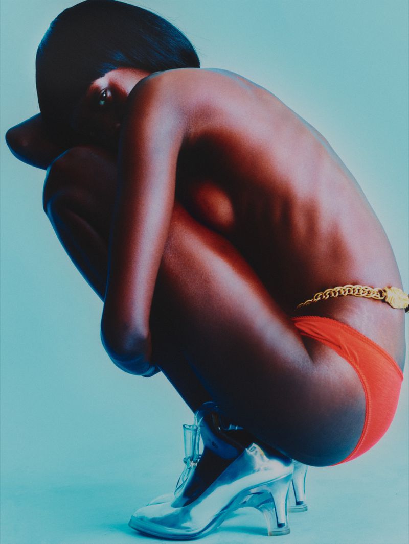 A dark-skinned female model crouches with her head resting on her knees. She is wearing red underwear and shiny silver high heels.