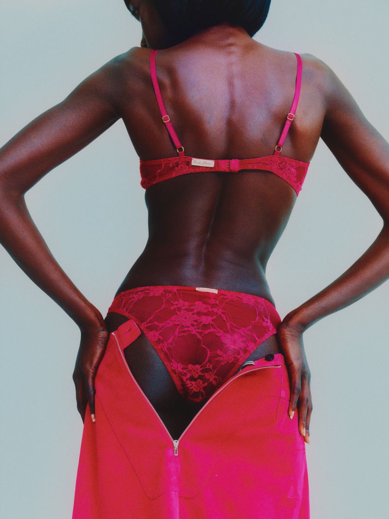 Rear view of a slender Black woman in red lace lingerie and a bright pink unzipped skirt, posing against a light background.