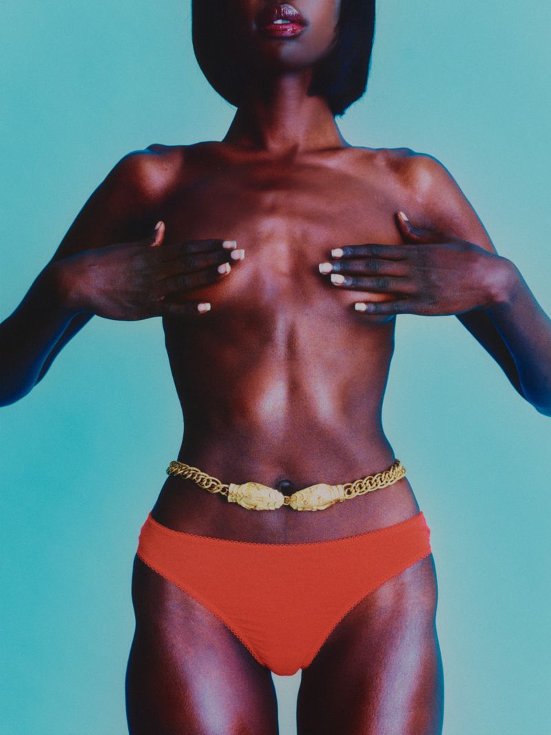 Close-up studio shot of a Black woman wearing a gold chain belt and orange bikini bottoms against a pale blue background.