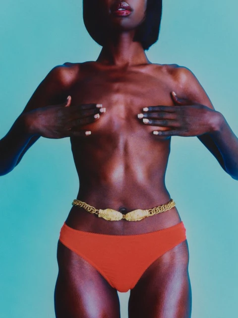 Close-up studio shot of a Black woman wearing a gold chain belt and orange bikini bottoms against a pale blue background.