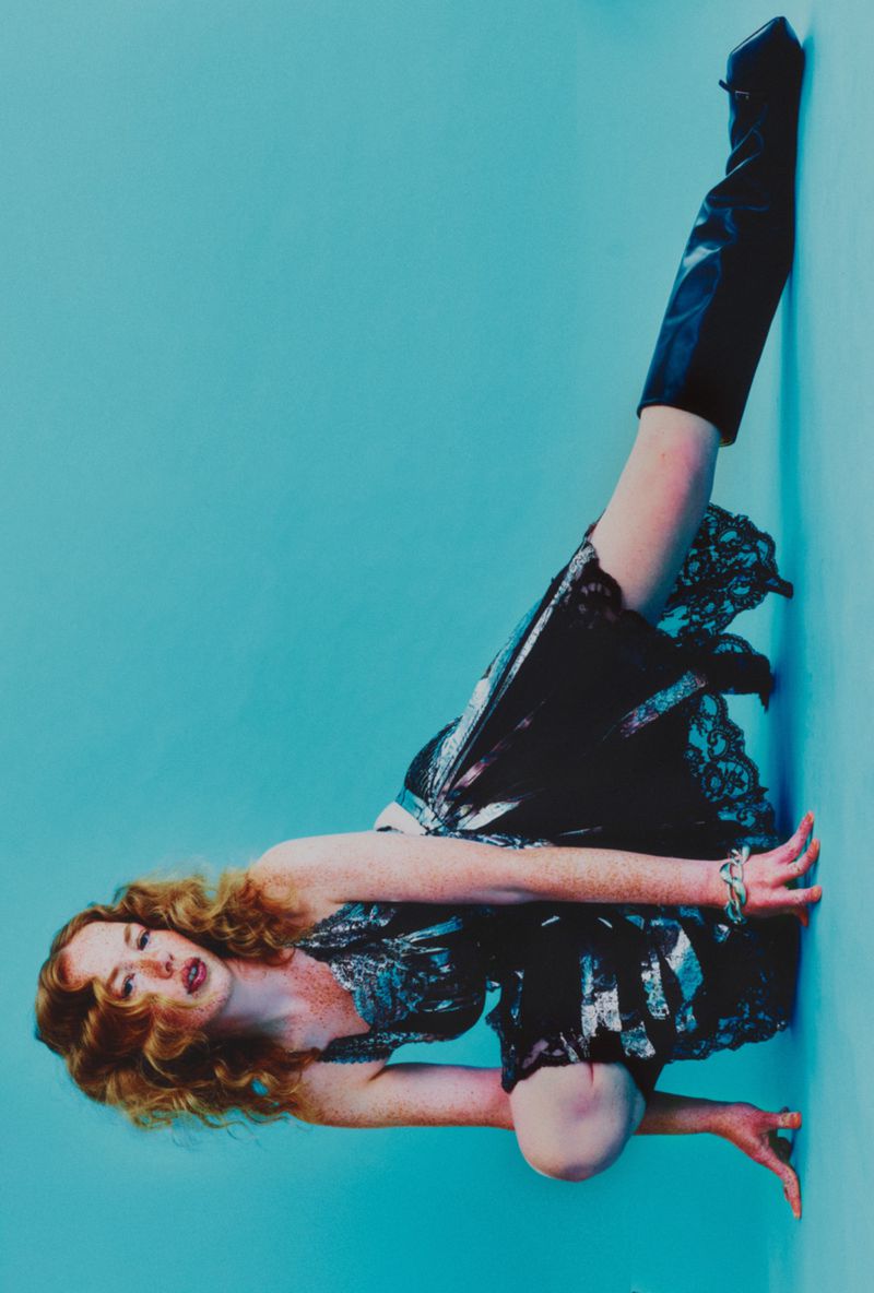 Adult female with red hair in black lace dress and thigh-high boots poses in a non-standing position against a blue wall.