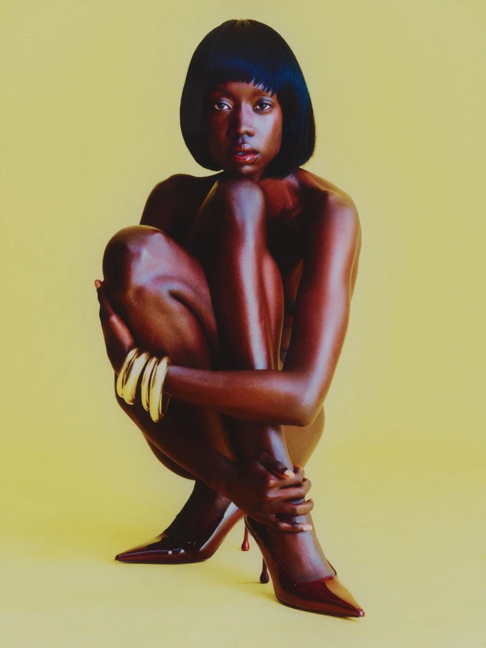 Black woman with a sleek black bob crouching against a yellow background wearing gold bangles and glossy pointed high heels.