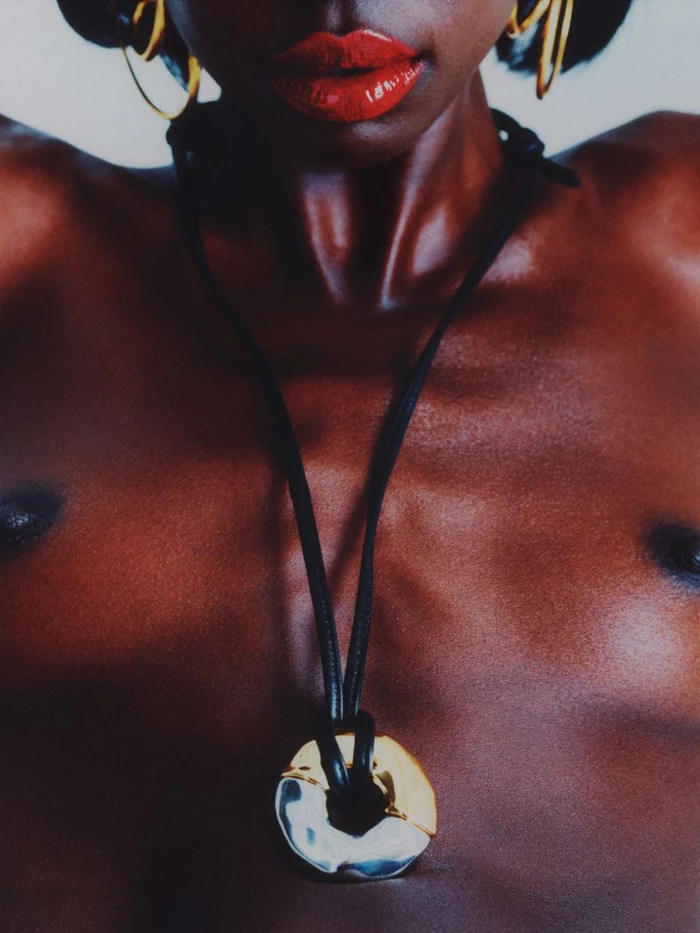 Close-up portrait of a black woman wearing glossy red lipstick, gold hoop earrings, and a pendant necklace against a light background