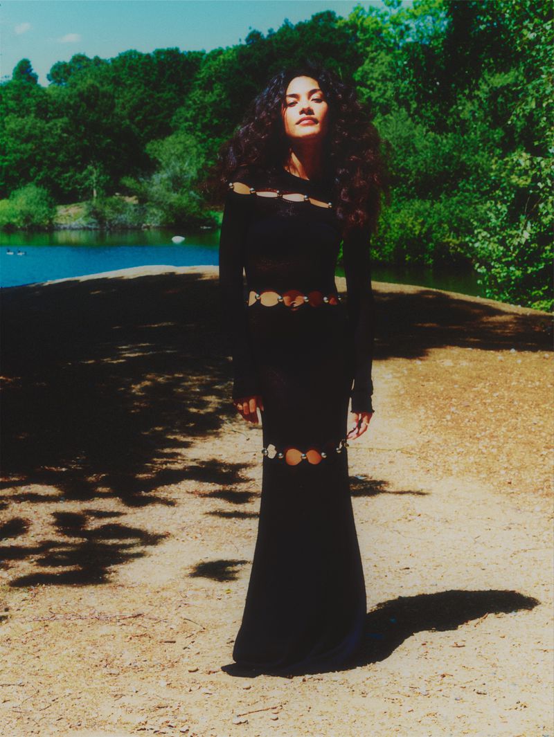 A woman in a long black dress with cutouts poses on a path with a lake and green trees in the background on a sunny day.