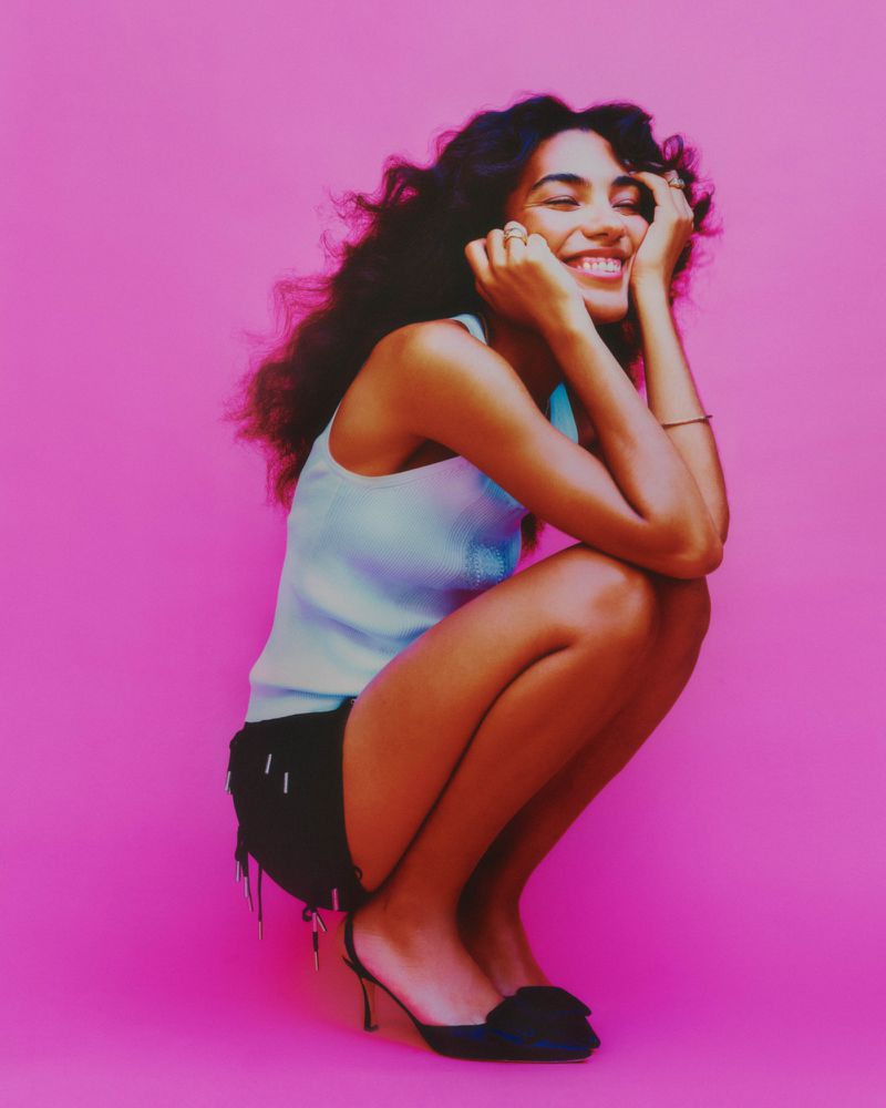 Smiling woman with dark curly hair, wearing a light blue tank top and black shorts, posing against a bright pink background in a studio.