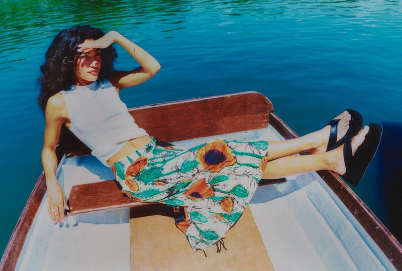 Woman in a boat on the water shielding her eyes from the sun, wearing a floral skirt, tank top, and sandals. Sunny day on the lake.