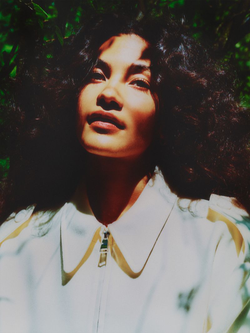 Portrait of a woman with dark curly hair looking up, wearing a white jacket with gold trim, surrounded by green foliage.