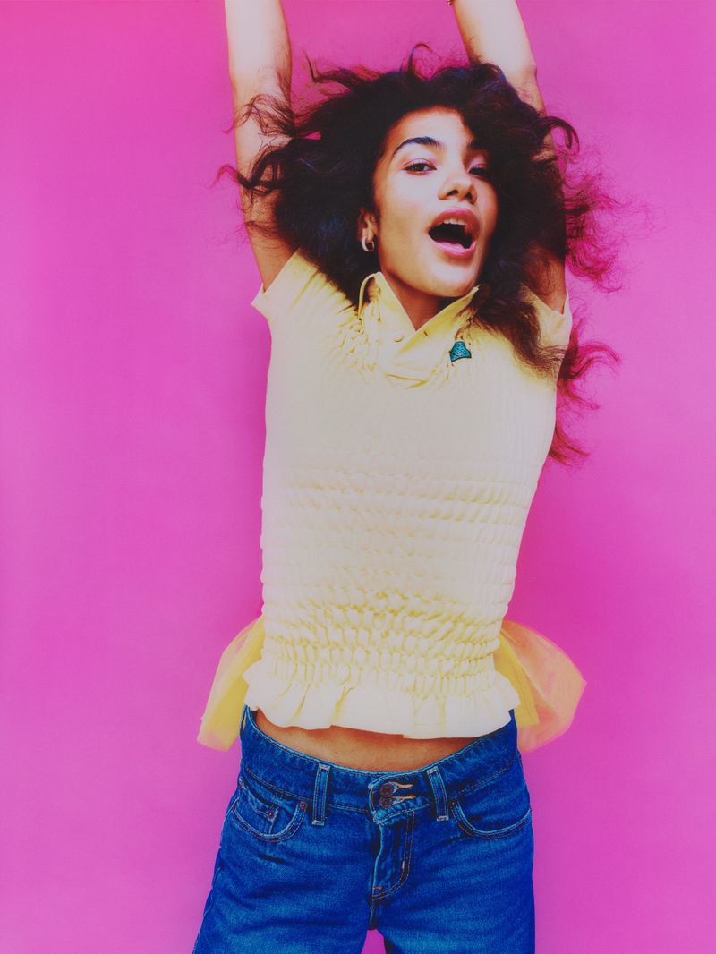 A young woman with curly brown hair is joyfully jumping with her arms raised against a bright pink background, wearing a yellow top and jeans.
