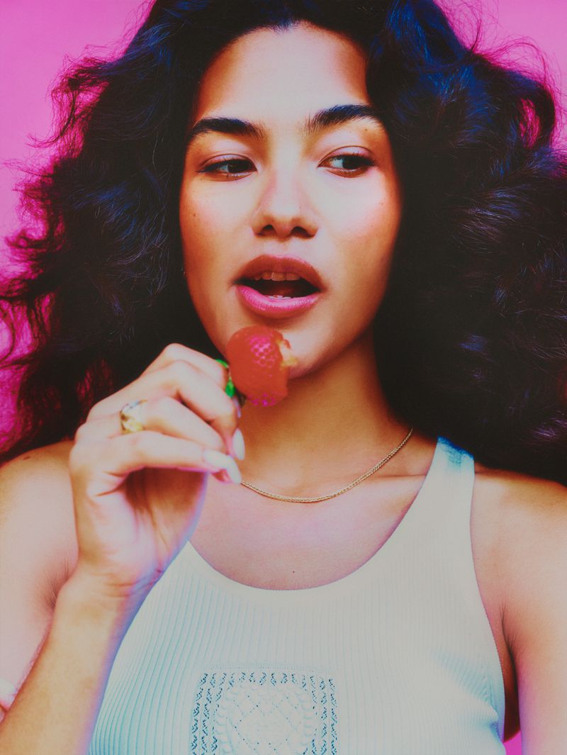 Portrait of an Asian woman with dark curly hair eating a strawberry against a pink background, wearing a white tank top.