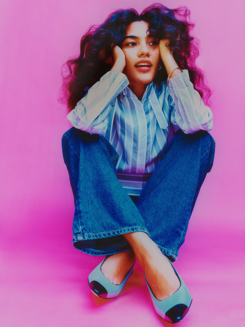 Young woman poses in casual clothes with blue jeans and striped shirt against a pink background in a studio setting.