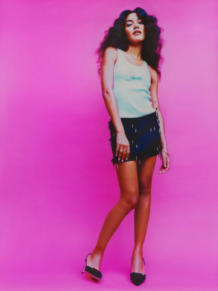 Singer Olivia Dean poses full-length against a vivid pink studio backdrop wearing a pale blue tank top and embellished black mini skirt.