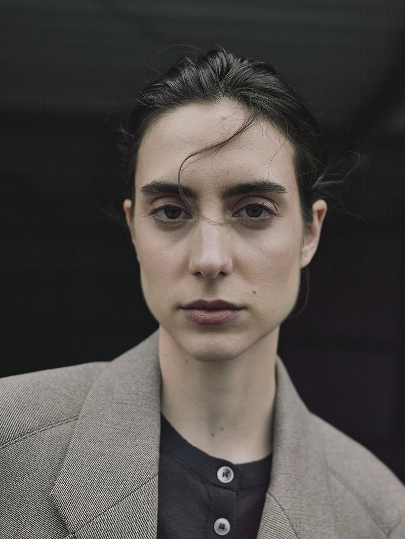 Portrait of a young woman with dark hair and brown eyes wearing a blazer against a dark background in a medium shot.